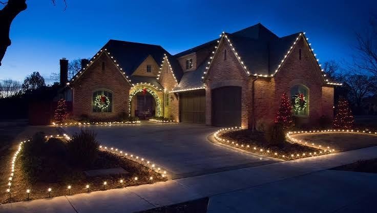 Brick mansion decorated with warm white Christmas lights outlining the roofline, driveway, and landscaping at dusk