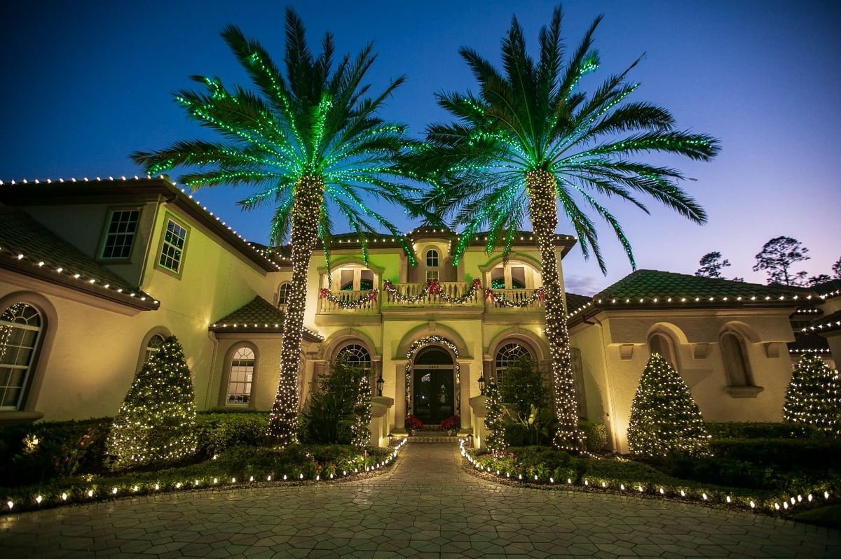 Luxury home illuminated at night with palm trees, holiday lights along pathway, and yellow Mediterranean-style architecture